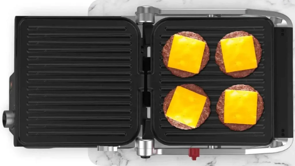 Grill Elétrico Multiuso Oster 2 em 1 aberto em 180 graus funcionando como chapa, preparando quatro hambúrgueres com queijo simultaneamente