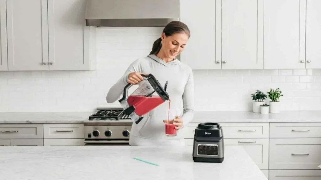 Mulher despejando um smoothie vermelho da jarra do Liquidificador Ninja Standalone Professional Plus em um copo na cozinha branca