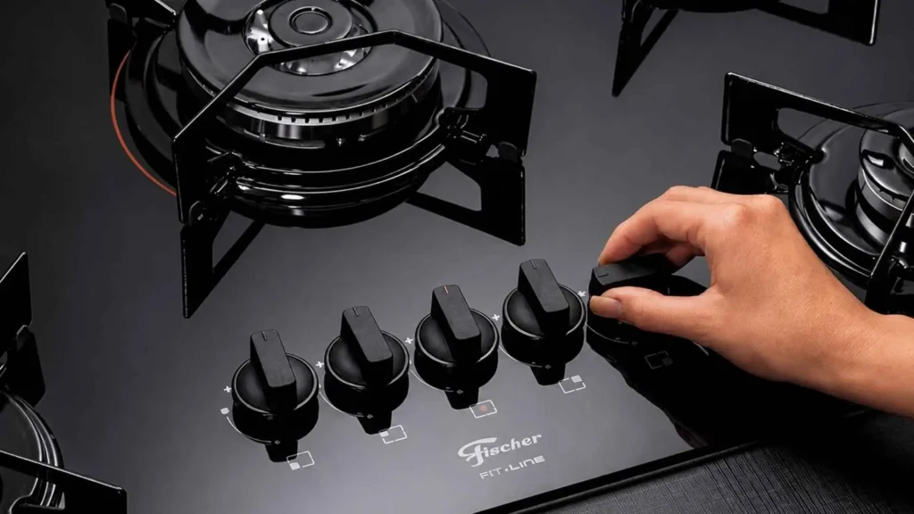 Mão ajustando o botão de controle e acendimento superautomático no cooktop a gás Fischer 5 bocas Fit Line com mesa de vidro preto