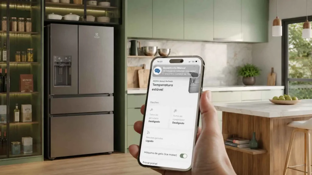 Mão segurando smartphone com o aplicativo aberto controlando as funções inteligentes da Geladeira Electrolux Expert IM9CS com o eletrodoméstico ao fundo