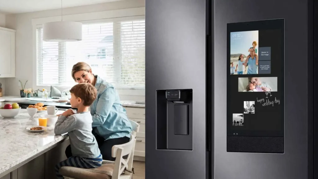 Detalhe do painel interativo da Geladeira Samsung RS58 585L mostrando fotos e aplicativos ao lado de uma família na cozinha